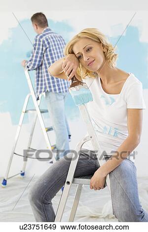 Tired woman holding paintbrush with man painting in background View Large Photo Image Stock Photo - Tired woman holding paintbrush with man painting in background. Fotosearch