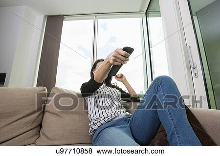 Stock Photo - Woman changing channels while watching TV on sofa at home. Fotosearch