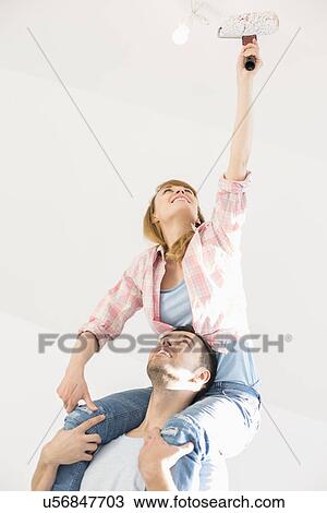 Stock Image - Woman on man's shoulders painting ceiling with paint roller. Fotosearch