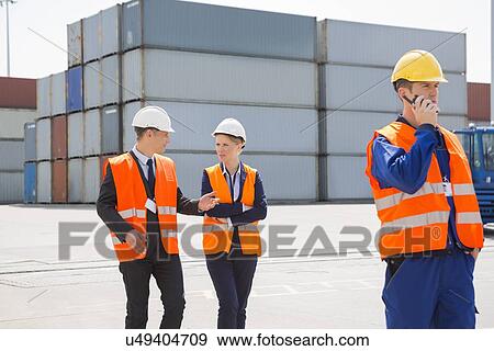 Stock Photo - Worker using walkie-talkie while colleagues discussing in shipping yard. Fotosearch