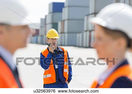 Stock Photograph - Worker using walkie-talkie while colleagues discussing in foreground at shipping yard. Fotosearch