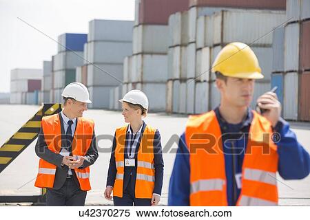 Stock Photography - Worker using walkie-talkie while colleagues discussing in shipping yard. Fotosearch