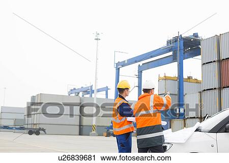 Stock Image - Workers discussing over blueprint in shipping yard. Fotosearch