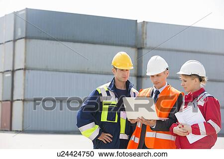 Stock Image - Workers discussing over laptop in shipping yard. Fotosearch