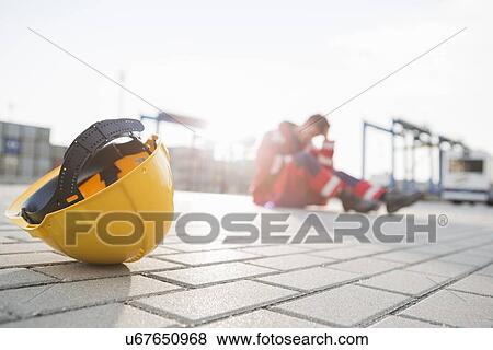 Stock Photo - Yellow hardhat at shipyard with depressed male worker in background. Fotosearch