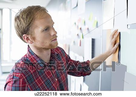 Young businessman reading documents on wall at creative office View Large Photo Image Stock Photo - Young businessman reading documents on wall at creative office. Fotosearch