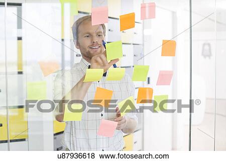 Stock Photo - Young businessman reading sticky paper on glass wall at creative office. Fotosearch