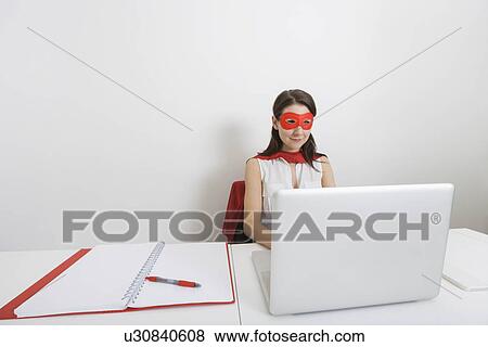 Young businesswoman dressed as superhero using laptop at desk in office View Large Photo Image Stock Photo - Young businesswoman dressed as superhero using laptop at desk in office. Fotosearch
