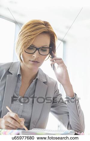 Stock Image - Young businesswoman on call while writing at desk in office. Fotosearch