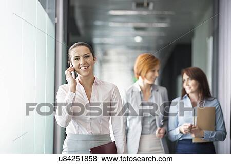 Stock Photo - Young businesswoman on call with colleagues in background at office corridor. Fotosearch