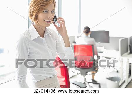 Young businesswoman talking on cell phone with colleagues in background at office View Large Photo Image Stock Photograph - Young businesswoman talking on cell phone with colleagues in background at office. Fotosearch