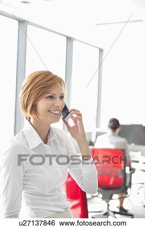 Stock Photograph - Young businesswoman talking on cell phone with colleagues in background at office. Fotosearch