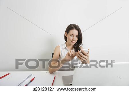 Stock Photo - Young businesswoman text messaging through cell phone at desk in office. Fotosearch