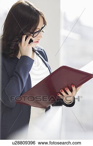 Young businesswoman using cell phone while reading file in office View Large Photo Image Stock Photograph - Young businesswoman using cell phone while reading file in office. Fotosearch