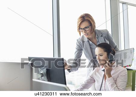 Stock Image - Young businesswoman using landline phone while colleague pointing at computer monitor in office. Fotosearch