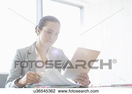 Young businesswoman using tablet PC while reading book in office View Large Photo Image Stock Image - Young businesswoman using tablet PC while reading book in office. Fotosearch