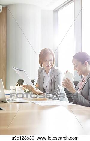 Stock Image - Young businesswomen having lunch at table in office. Fotosearch