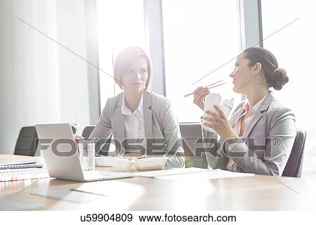 Stock Photo - Young businesswomen having lunch at table in office. Fotosearch
