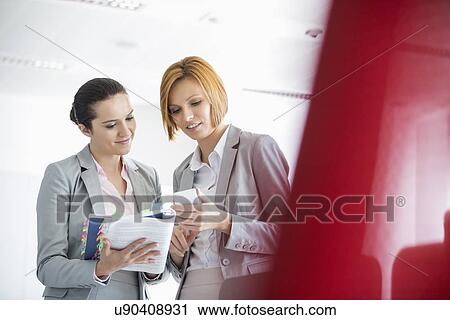 Stock Image - Young businesswomen with book while using cell phone in office. Fotosearch