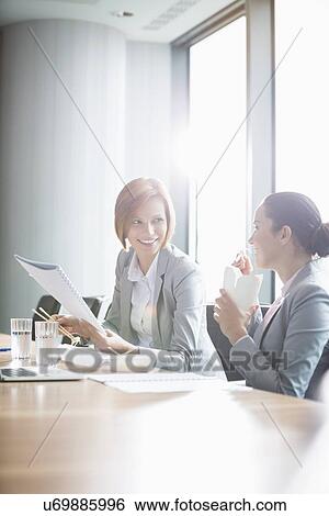 Young businesswomen working while having lunch at table in office View Large Photo Image Stock Photograph - Young businesswomen working while having lunch at table in office. Fotosearch