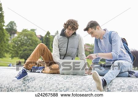 Young male college friends with laptop studying together in park View Large Photo Image Stock Photo - Young male college friends with laptop studying together in park. Fotosearch