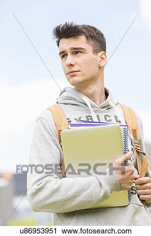 Young male college student holding books while looking away outdoors View Large Photo Image Stock Image - Young male college student holding books while looking away outdoors. Fotosearch