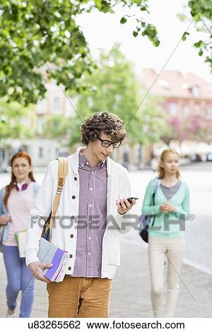 Stock Image - Young male student using cell phone with friends in background on street. Fotosearch