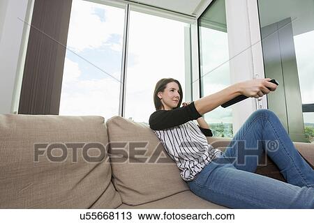 Stock Image - Young woman watching TV on sofa at home. Fotosearch