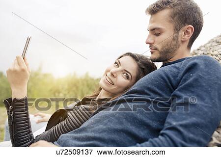 Stock Photo - Attractive young couple leaning back looking at smartphone. Fotosearch
