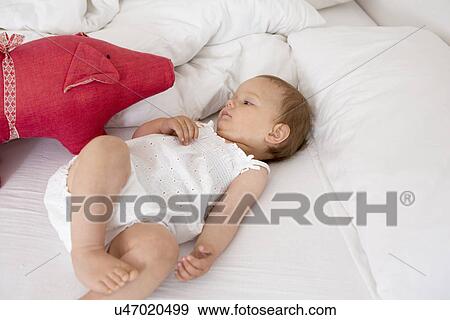 Baby girl lying on bed looking at soft toy View Large Photo Image Stock Photo - Baby girl lying on bed looking at soft toy. Fotosearch