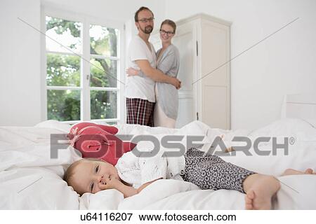 Baby girl lying on bed, parents in background View Large Photo Image Stock Photo - Baby girl lying on bed, parents in background. Fotosearch