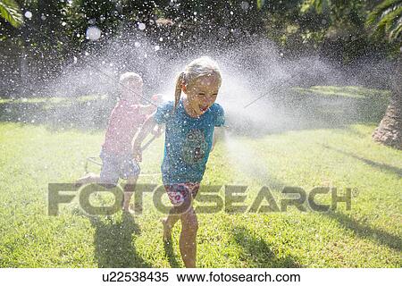 Stock Photography - Boy running after girl in garden with water sprinkler. Fotosearch