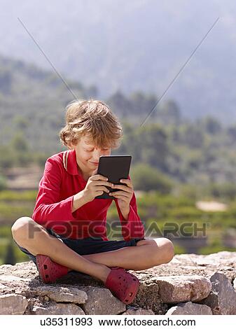 Stock Image - Boy sitting on stone wall looking at digital tablet, Majorca, Spain. Fotosearch