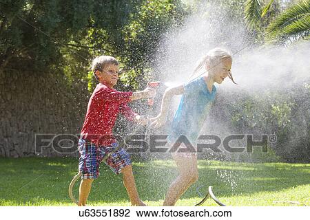 Stock Image - Boy splashing girl in garden with water sprinkler. Fotosearch