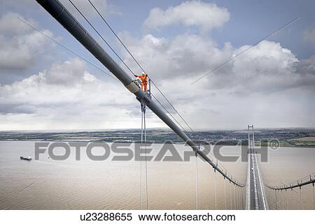 Stock Photography - Bridge worker walking on cable of suspension bridge. The Humber Bridge, UK was built in 1981 and at the time was the world's largest single-span suspension bridge. Fotosearch