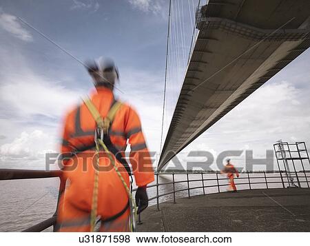 Stock Photo - Bridge workers walking under suspension bridge. The Humber Bridge, UK was built in 1981 and at the time was the world's largest single-span suspension bridge. Fotosearch