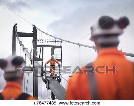 Stock Photography - Bridge workers watching coworker on cable of suspension bridge. The Humber Bridge, UK was built in 1981 and at the time was the world's largest single-span suspension bridge. Fotosearch