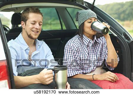 Stock Image - Couple sitting at rear of car with legs tucked in sleeping bag. Fotosearch