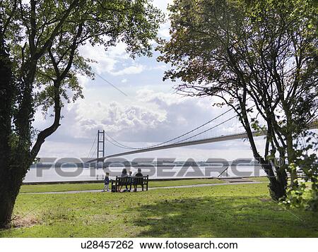 Stock Image - Family sitting together on bench looking at suspension bridge. The Humber Bridge, UK was built in 1981 and at the time was the world's largest single-span suspension bridge. Fotosearch