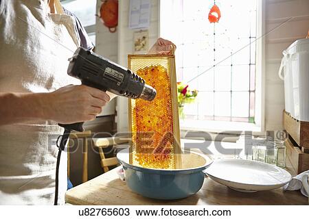 Stock Image - Female beekeeper in kitchen melting wax on frame of honey. Fotosearch