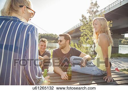 Stock Image - Four young friends drinking beer on riverside picnic bench. Fotosearch