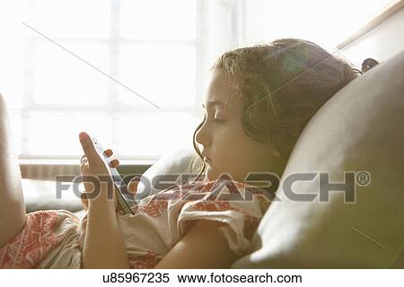 Stock Photography - Girl lying on living room sofa browsing smartphone. Fotosearch