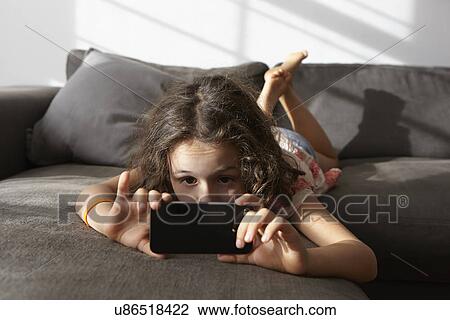 Stock Image - Girl lying on living room sofa looking at smartphone. Fotosearch
