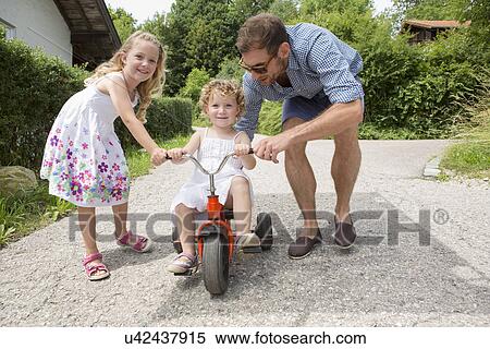 Stock Photography - Girl riding tricycle with father and sister. Fotosearch