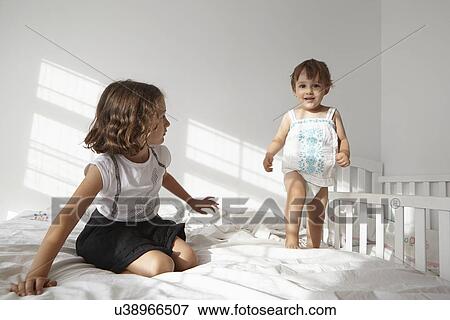 Stock Photo - Girl watching female toddler cousin to toddle on bed. Fotosearch