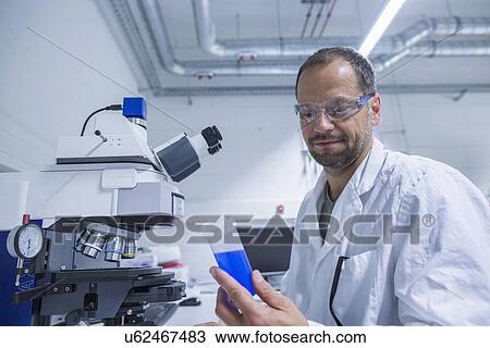Stock Image - Lab assistant looking at tube of blue liquid. Fotosearch