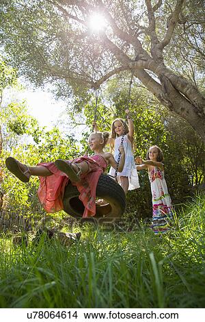 Picture - Low angle view of three girls playing on tree tire swing in garden. Fotosearch