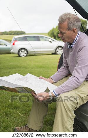 Stock Image - Man sitting on car boot reading map. Fotosearch