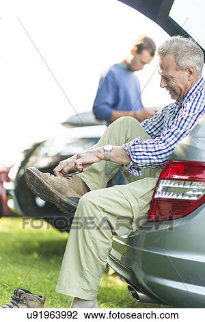 Stock Image - Man sitting on car boot tying shoelaces. Fotosearch