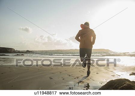 Stock Photo - Mature man running on sand, along coastline. Fotosearch